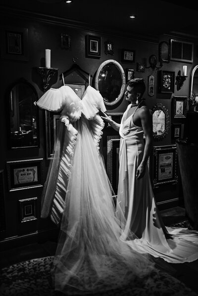 a black and white photo of a LGBTQ+  groom, holding  out his wedding dress