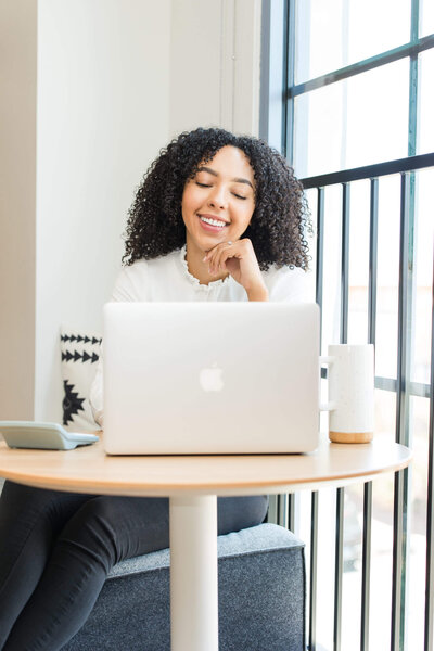Smiling woman with curly hair working on a laptop at a sunlit table by a window. She appears relaxed and content, with a mug and notebook nearby.