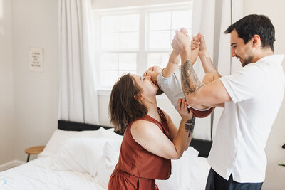 a father helps his toddler daughter stand on the bed in their brightly lit bedroom