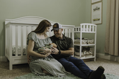 Verona B Photography captures an adorable sleeping newborn baby held lovingly by his parents as they smile at him. The family is on the floor of the nursery leaning up against the crib. Beautiful in-home session near Akron, Ohio. 