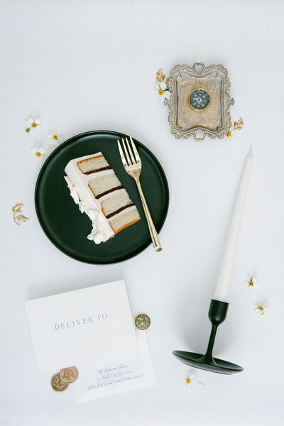 A refined flatlay featuring a slice of wedding cake, an elegant black candle, vintage coins, and delicate stationery at a luxury wedding at Graylyn Estate in Winston-Salem, North Carolina.