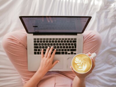 Photo from above of a laptop sitting on someone's lap in pink pants holding a mug of coffee