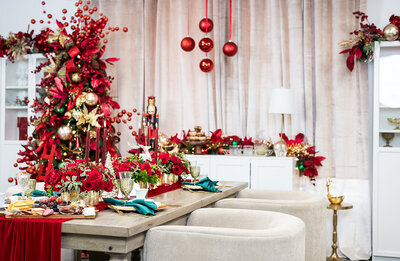 Elegant holiday tablescape with lush red floral centerpieces, gold-rimmed glassware, emerald green napkins, and velvet accents, set beside a lavish Christmas tree adorned with red and gold ornaments, designed by Wells Design Co.