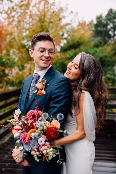 Bride and groom embracing outdoors in front of autumn trees, the bride holding a vibrant bouquet of poppies, cosmos, and ranunculus in warm, saturated tones.