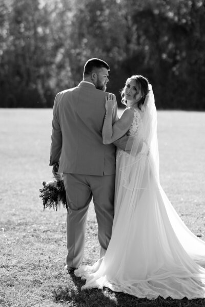 A stunning bride looks back over her shoulder at the camera in a striking black and white photo as her arm rests on her new husband,