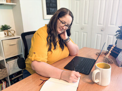 Ali wearing yellow shirt journaling at desk with laptop, mug, and creative decor