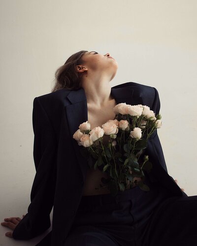 Brunette woman sitting on floor with head back in a black blazer with fresh flowers on chest