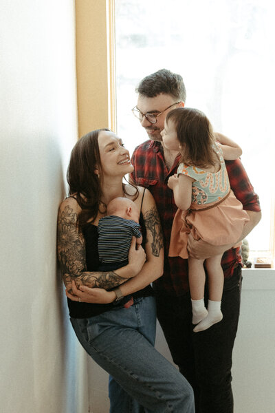 A family of four in matching plaid shirts sits together in front of a window in their home.