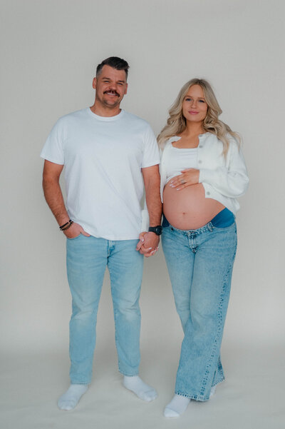 Studio photo session. Couple poses in front of a white backdrop. Couple holds hands while the woman's other hand is placed on top of her belly.