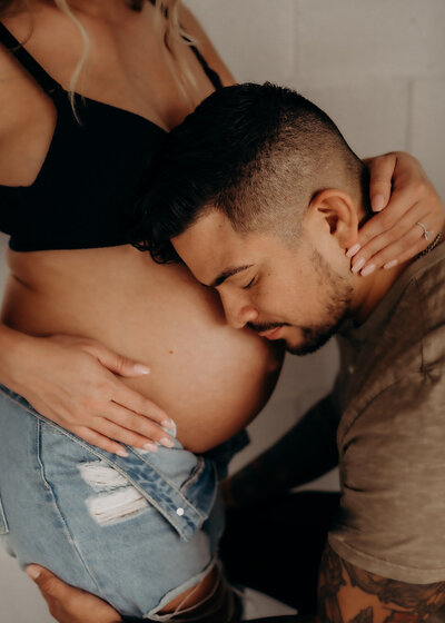A very handsome husband kisses his wife's pregnant belly in a close-up image where she holds his neck while wearing a black bra and oversized denim jeans.