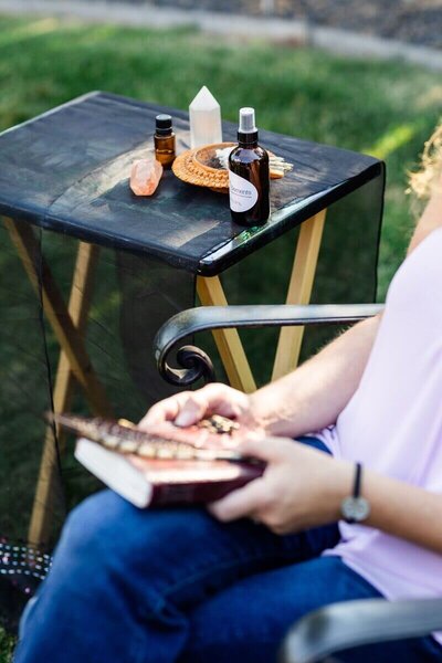 A woman sits with a journal on her lap next to a small outdoor table displaying healing tools including crystals, essential oils, a spray bottle, and a bundle of sage.