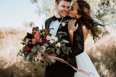Bride kisses groom’s cheek while holding colorful bouquet at a sunny Breckenridge wedding.