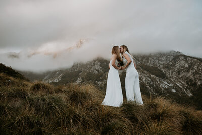 A lesbian couple holds hands facing each other, kissing under a mountain peak in Slovenia. They are both wearing white wedding attire.