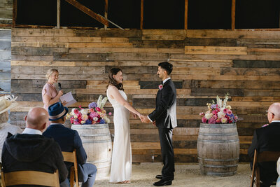 Bride and groom holding hands during a rustic indoor wedding ceremony, with a wooden plank backdrop, floral arrangements on wine barrels, and a woman officiating the ceremony while guests look on