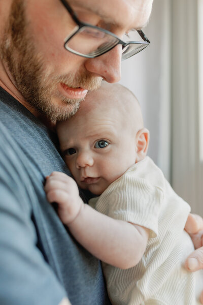 A tender father-child moment as a dad holds his newborn close to his chest, both bathed in soft natural light from a nearby window. The baby rests peacefully against him, exuding warmth and security. This intimate portrait highlights the deep connection between parent and child, a hallmark of KCP Families' emotive storytelling photography.
