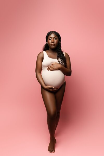 black pregnant mom posing for maternity pictures in pink body suit on pink backdrop