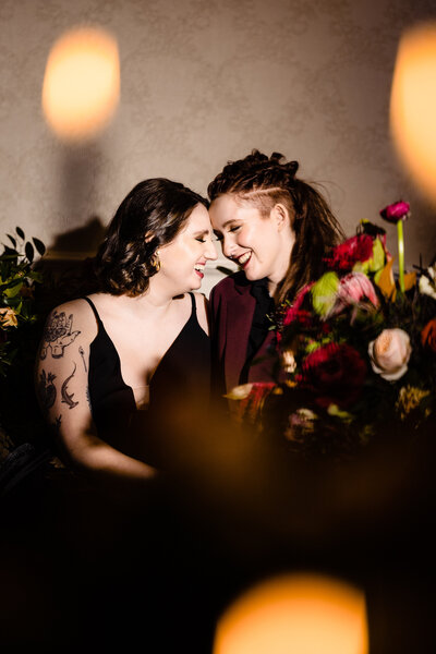 Two brides smiling with their foreheads touching surrounded by gothic-inspired wedding flowers