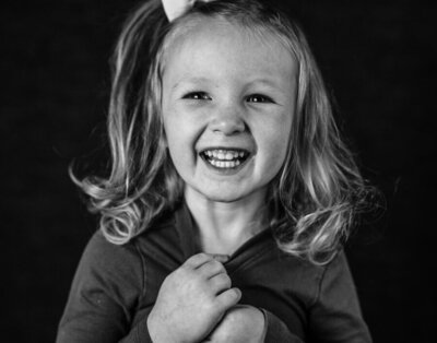 A preschool girl giggles and smiles as she  plays with her shirt during a school photo day that captures authentic child personalities and expressions.