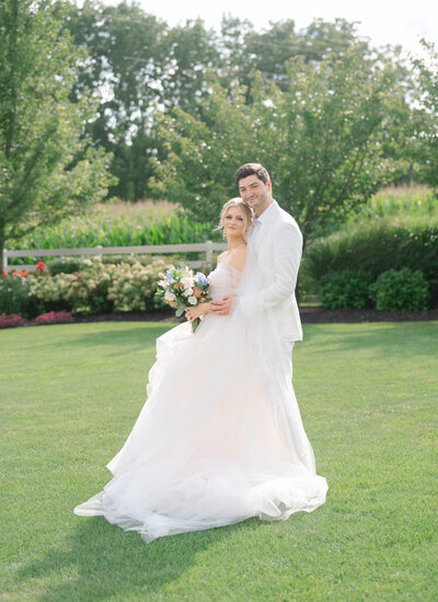 A bride and groom in a white suit smile at their summer wedding near a garden