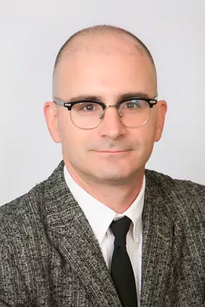 Headshot of a bald man with brown eyes, glasses, and a tweed blazer.