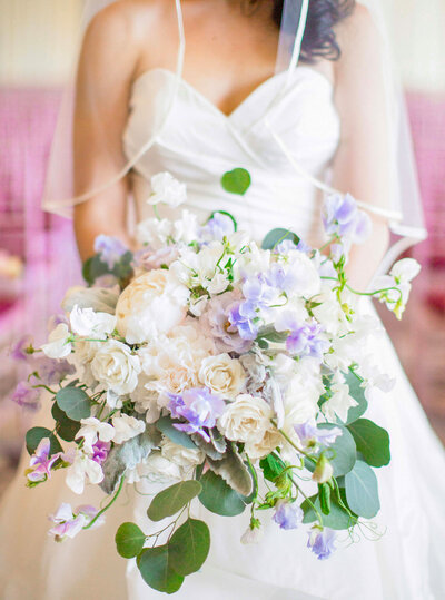A bride in a white dress holds a bouquet of white and lavender flowers with green leaves. The bouquet features roses and delicate blossoms, creating a soft, romantic arrangement. The brides veil is partially visible.