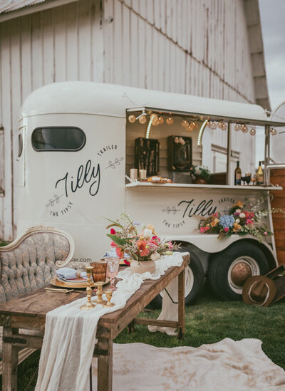 A vintage horse trailer bar, "Tilly," with floral decor and a rustic tablescape. Representing Idaho Tipsy Trailers vendor profile for the Crush Collective wedding show.