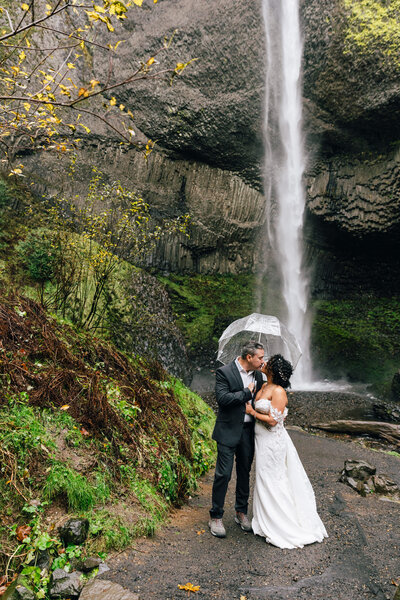 A couple kisses under a clear umbrella at the base of a waterfall in Oregon.