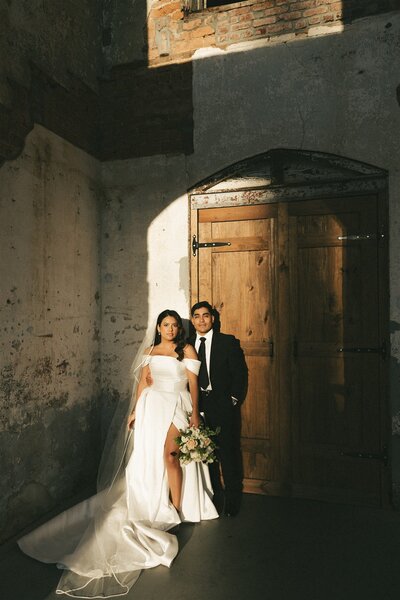 Newlyweds smile together in a rustic room with large wooden door