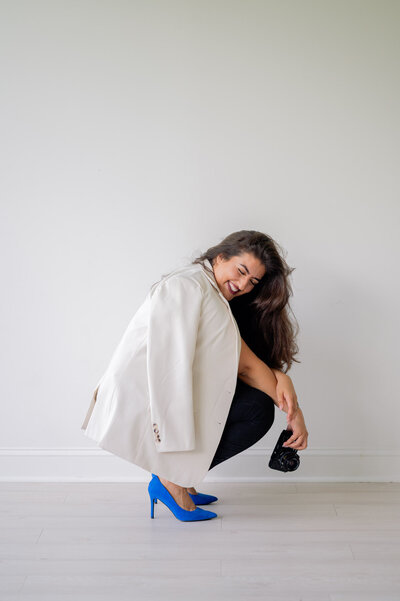 Hiba is wearing a white jacket draped around her shoulders and bright blue high heels while kneeling close to the ground. She's in an all white studio.