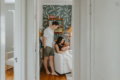 Mom sitting down holding newborn son with dad standing looking at baby