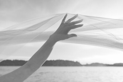 Best Rhode Island Wedding Photographers | Kelsey Sheehan Photography Timeless Rhode Island Weddings | A hand reaches up, gently holding translucent fabric against a light sky. In the background, a silhouette of trees lines a calm body of water.