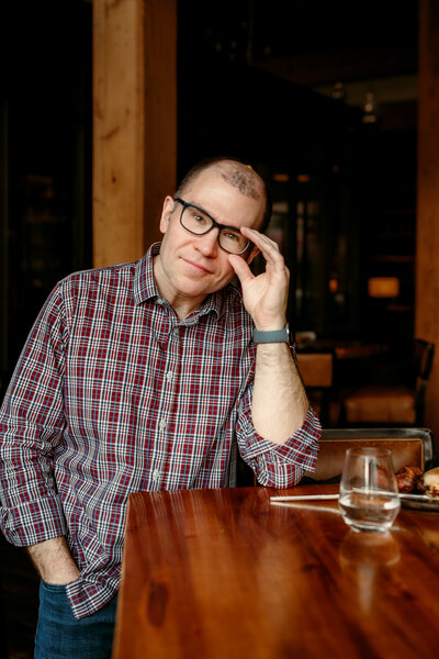 Candid dating profile photo of Ryan seated at a table with a drink, showcasing a thoughtful and approachable look for online dating apps.