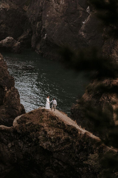 A boho couple with lanterns sitting on an outcropping with vast canyons in the background. The groom is wearing maroon. They look at each other and laugh.