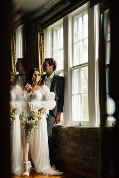 Bride and groom holding each other, gazing out of a window at their wedding venue, captured in soft, natural light for a romantic and intimate moment.