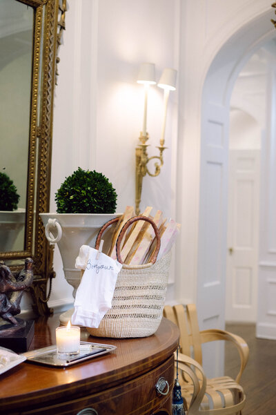 Woven market basket filled with baguettes, a ‘bonjour’ onesie, and a lit candle on an antique console under brass sconces.
