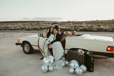2 brides sit in a vintage getaway car one wearing a tux and one wearing a white gown. There are disco balls outside the car of varying sizes . Photo taken by Amelia Island wedding photographer