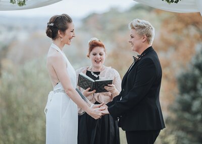 a brunette bride dressed in a white jumpsuit and a blonde bride dressed in a black suit recite their vows on a mountain top with their officiant