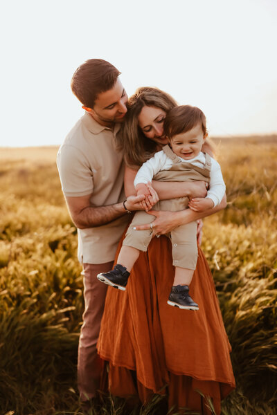 A father wrapping his arms around his wife and toddler boy in a golden field at sunset. 
