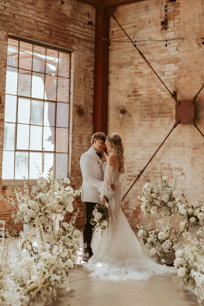 A bride and groom holding each other in a brick warehouse surrounded by gorgeous white floral installations. 