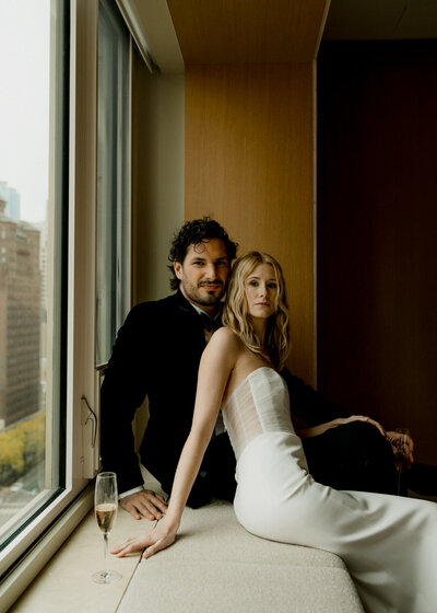 Blonde bride sits in strapless dress in front of brunette husband wearing a tux on a windowsill with a glass of champagne.