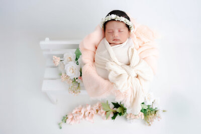 Baby swaddled in a bow wrap, laying on top of a bench prop with peach flowers and fabrics surrounding her. 
