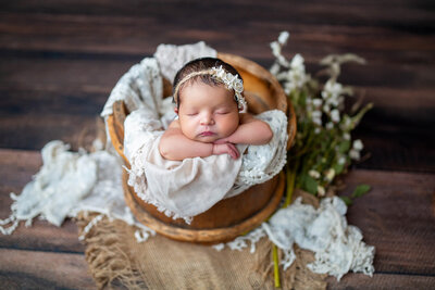Beautiful baby posed in a brown bucket with head resting on arms surrounded by lace and flowers. captured by Sarah Wyndham Photography