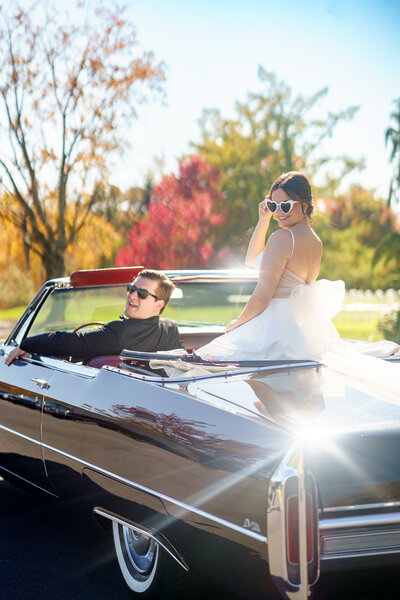 Groom driving convertible car while bride sits on the back wearing sunglasses