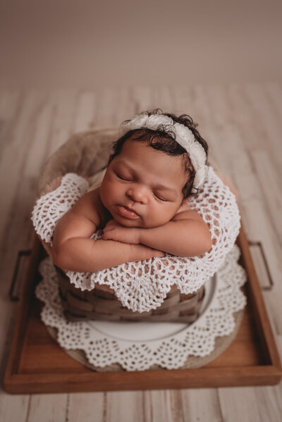 Newborn baby girl asleep and posed sitting in a basket with hands under her chin, wearing a white flower headband and white doily underneath of her