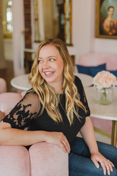 Smiling woman sitting in a pink chair at a cafe-style table with flowers.