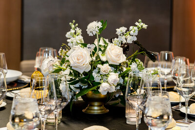 A lush white floral centerpiece in an elegant gold compote vase, featuring roses, stock, and delicate greenery. Surrounded by candlelight and glassware, this arrangement adds a timeless and sophisticated touch to the black-tie wedding table setting.