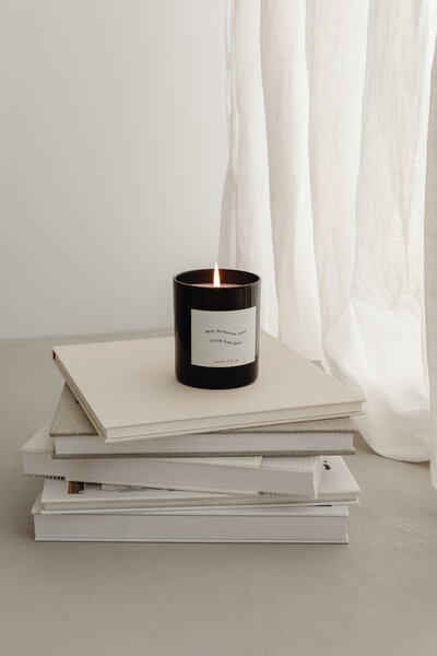 A lit candle in a sleek black jar sits atop a stack of neutral-colored books, set against soft white net curtains. The warm glow and minimalist styling create a tranquil atmosphere. Designed for The Artisan Candle’s social media campaign, this image embodies relaxation, home decor, and cozy living.