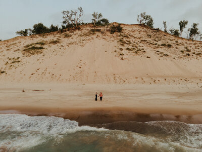 A couple standing hand in hand on the sandy shore at Indiana Dunes with soft waves and golden evening light.