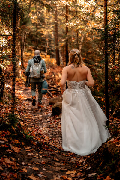 A couple hikes through the woods in their wedding attire from their sunrise elopement on top of a mountain in the adironacks in upstate ny