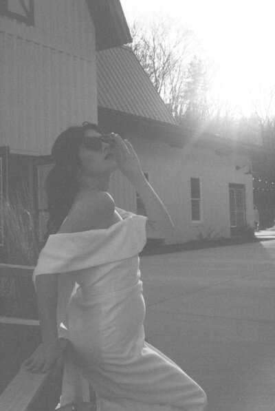 A black and white film photo of a modern bride in a short wedding dress and vintage sunglasses.